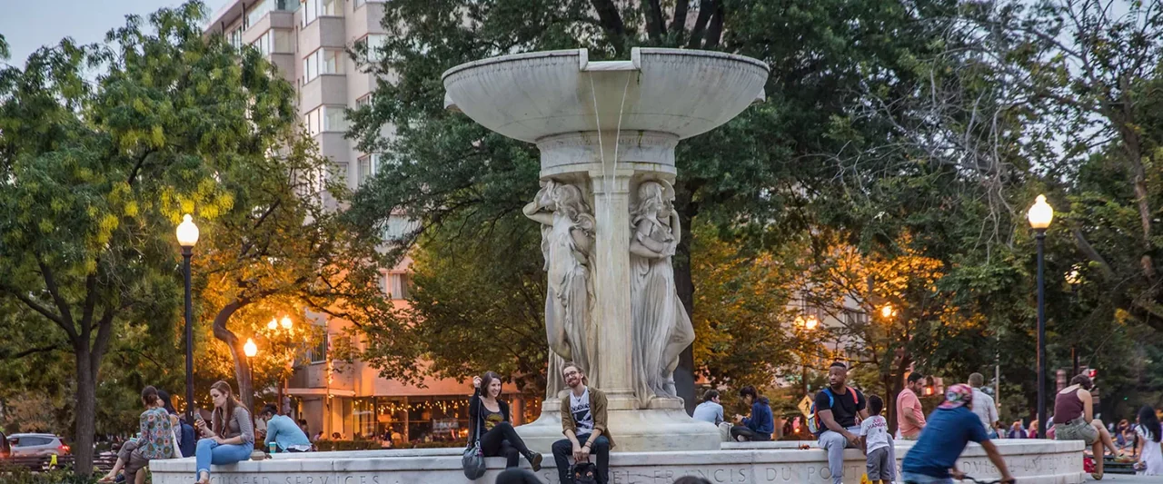 The marble fountain in Dupont Circle surrounded by trees and neighborhood buildings.