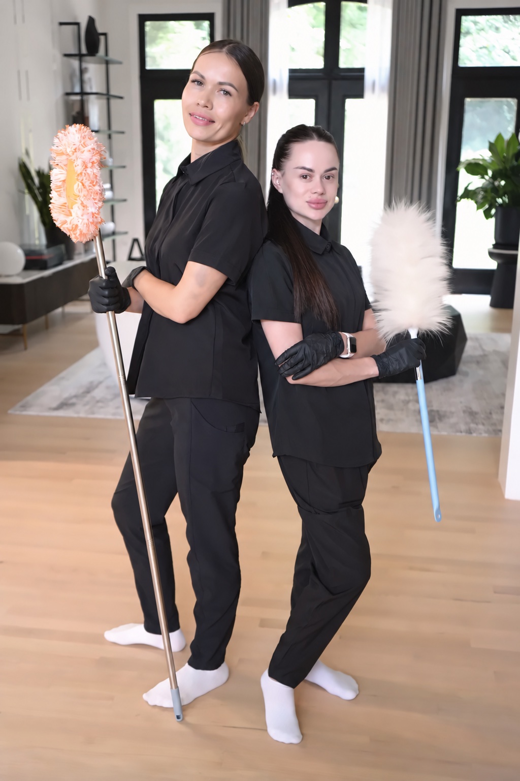 Two cleaners standing back to back holding dusters, in socks on a hardwood floor